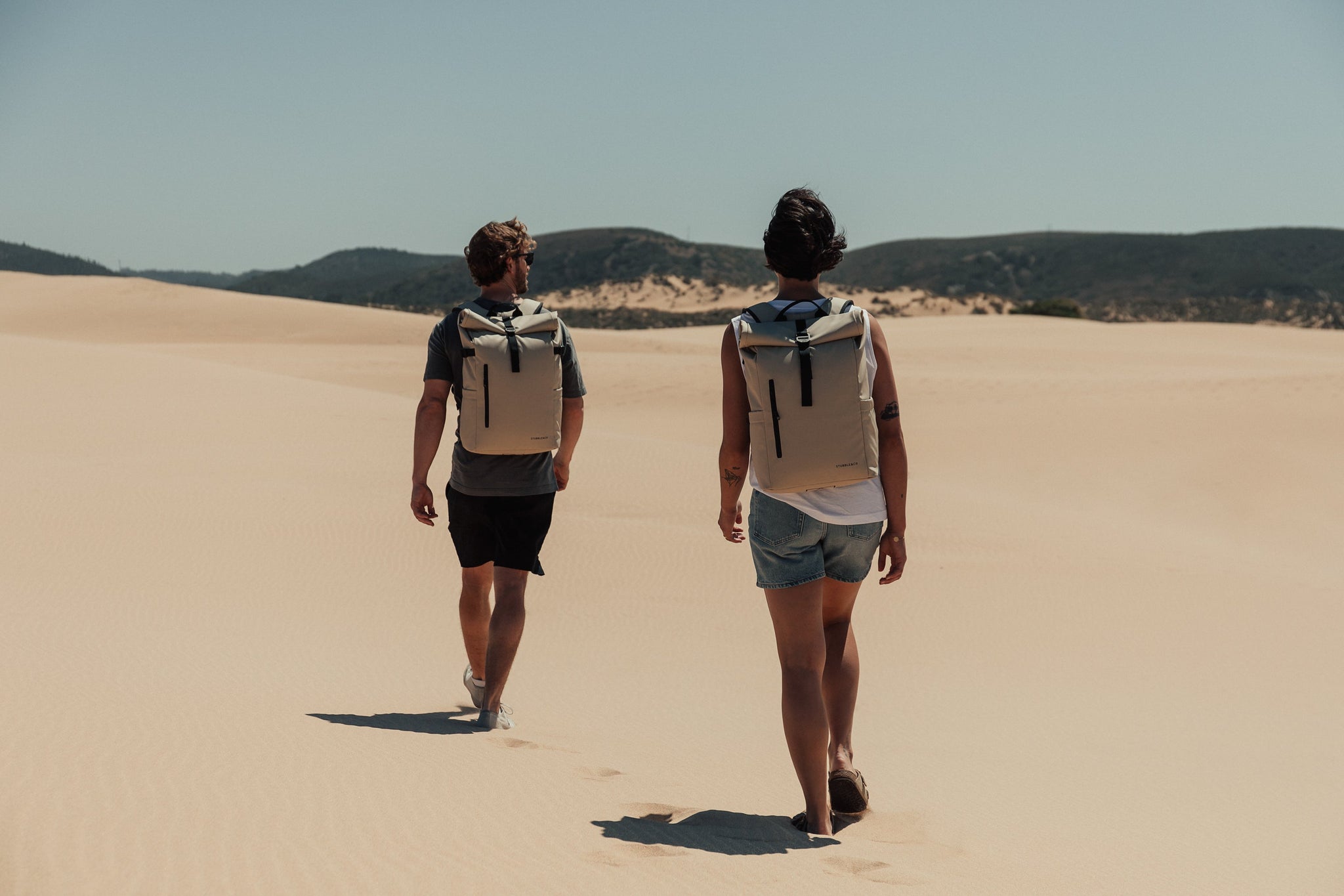 Man and women walking along beach wearing Roll Top backpacks