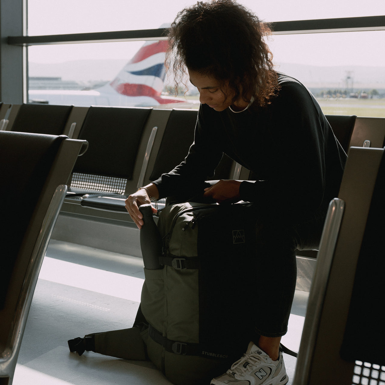A woman placing a bottle in the side bottle pcoket on an Urban Green Adventure Bag whilst she sits in an airport.
