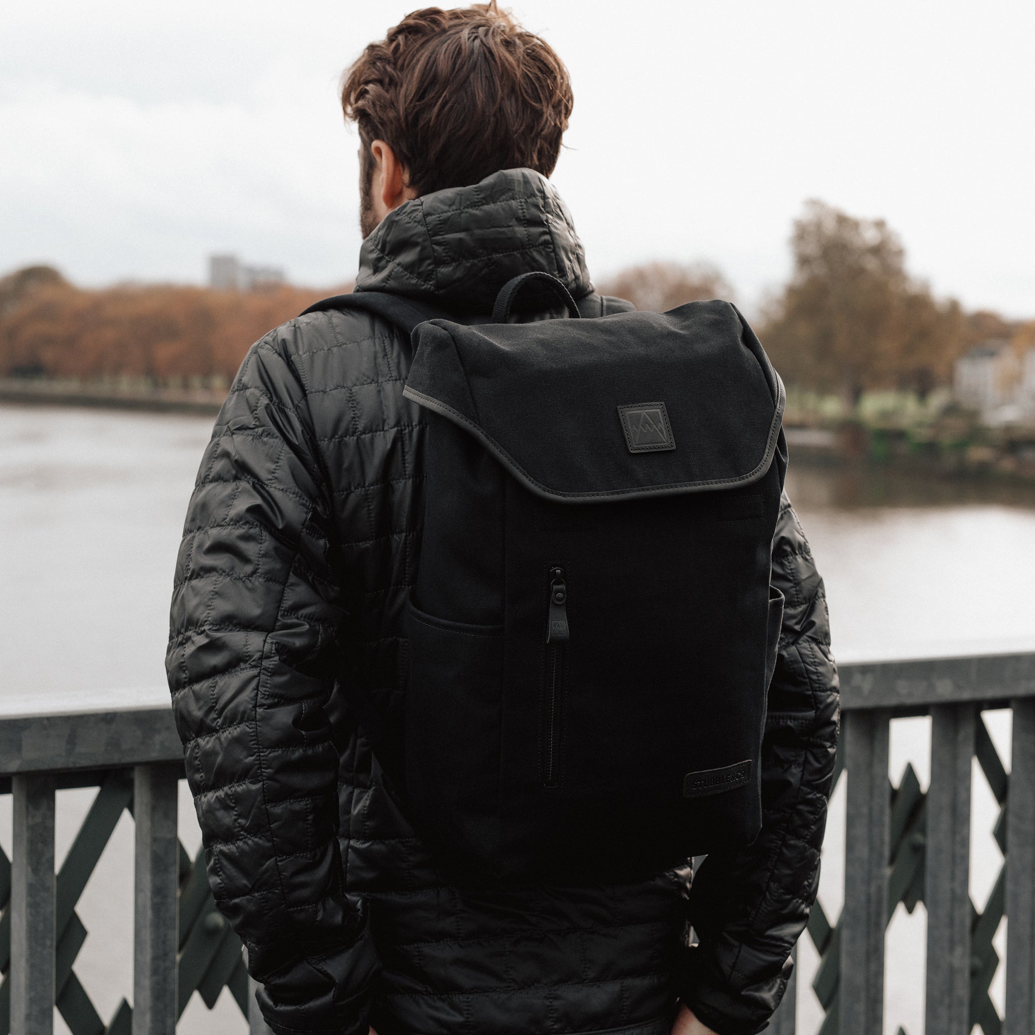 A man standing on a bridge wearing an All Black Daypack Outlet on his back