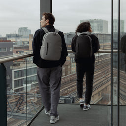 A man and woman wearing Everyday Backpack15L on their backs in black and Concrete