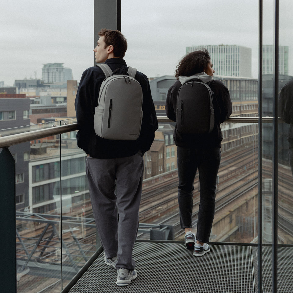 A man and woman wearing Everyday Backpack15L on their backs in black and Concrete