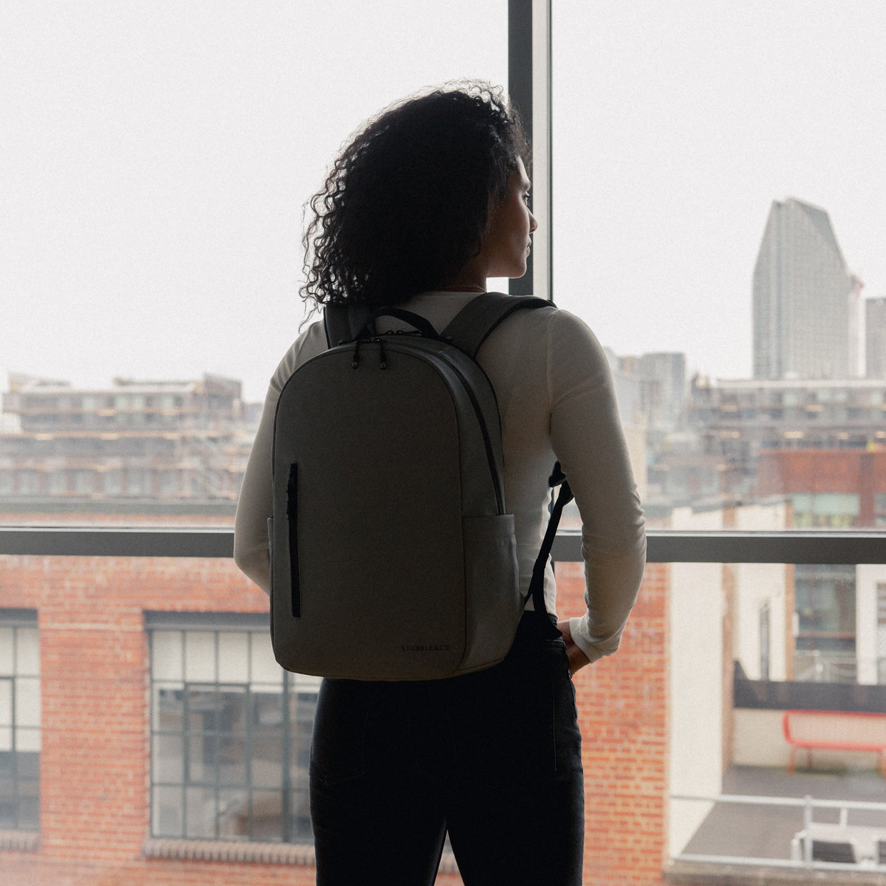 A woman looking out the window wearing an Everyday Backpack 15L in Concrete on her back