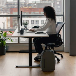 A woman sitting at a desk with an Everyday Backpack 15L in Concrete at her side