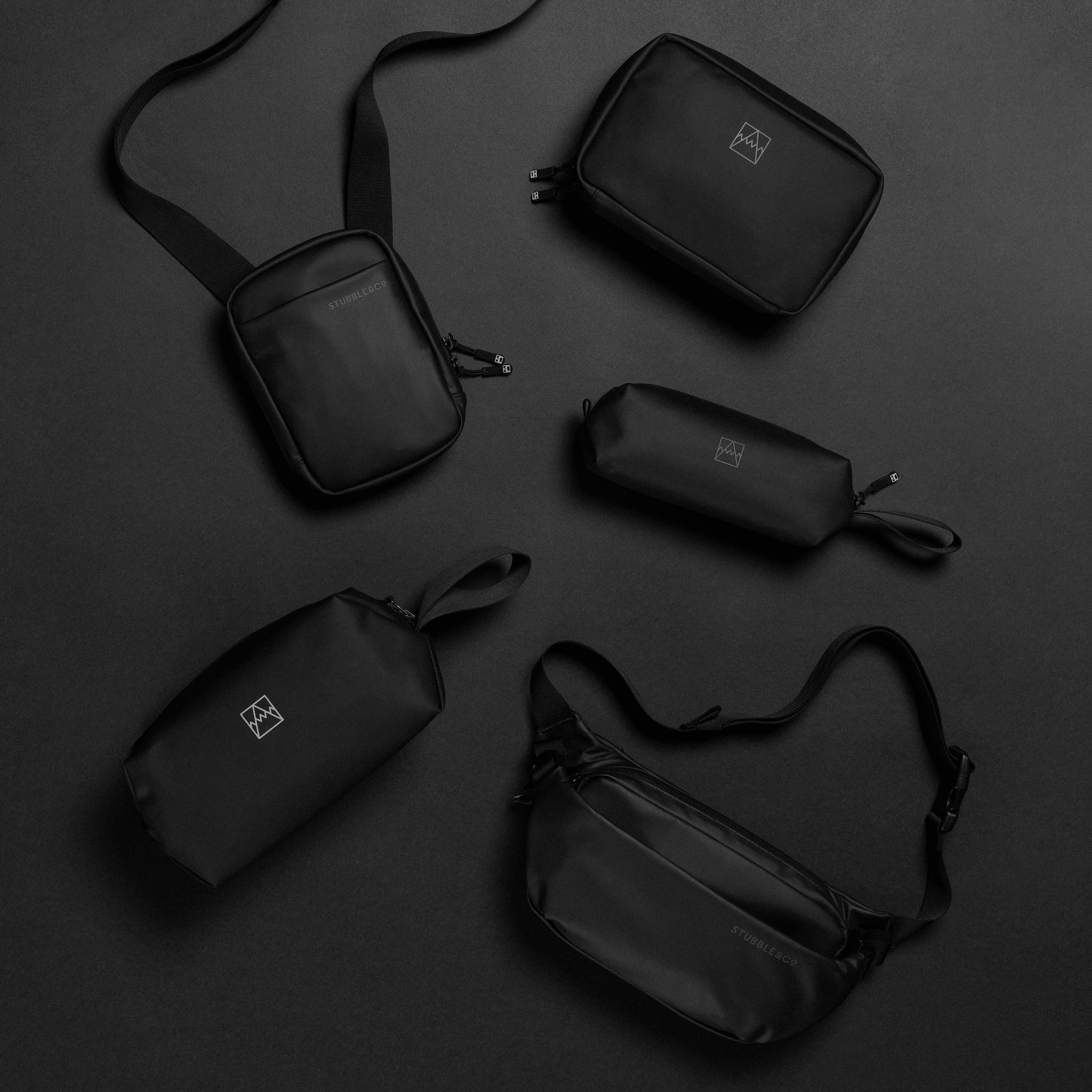 A studio shot of black shoulder bag, tech bag, pencil case, wash bag and crossbody bag lying on the floor