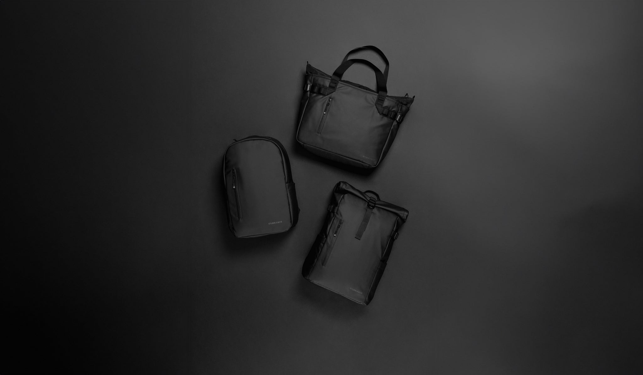 A studio shot of the tote bag, everyday backpack and roll top 20l in black lying on floor