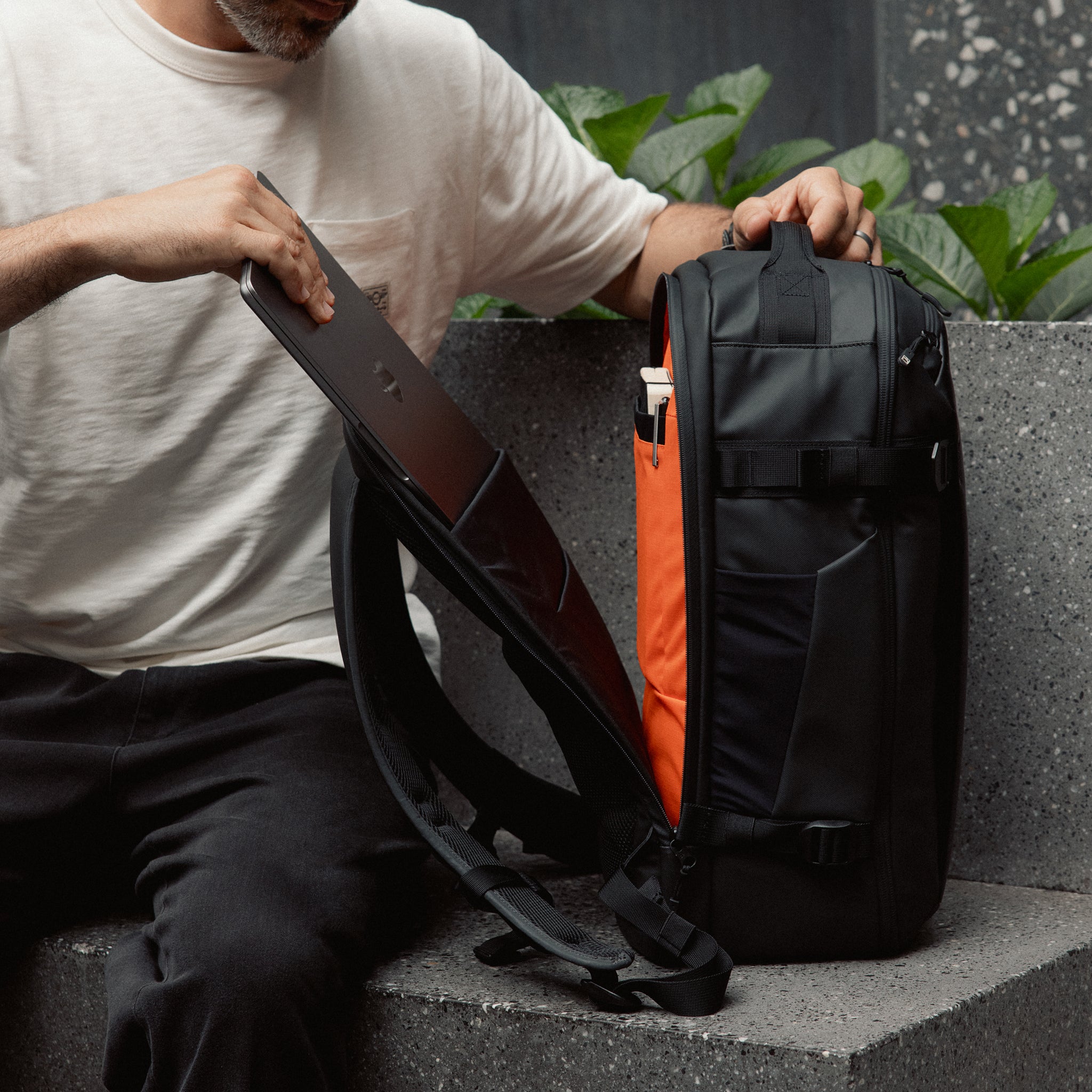 A lifestyle shot of a man placing a laptop into the laptop compartment in the  Hybrid Backpack 30L in All Black