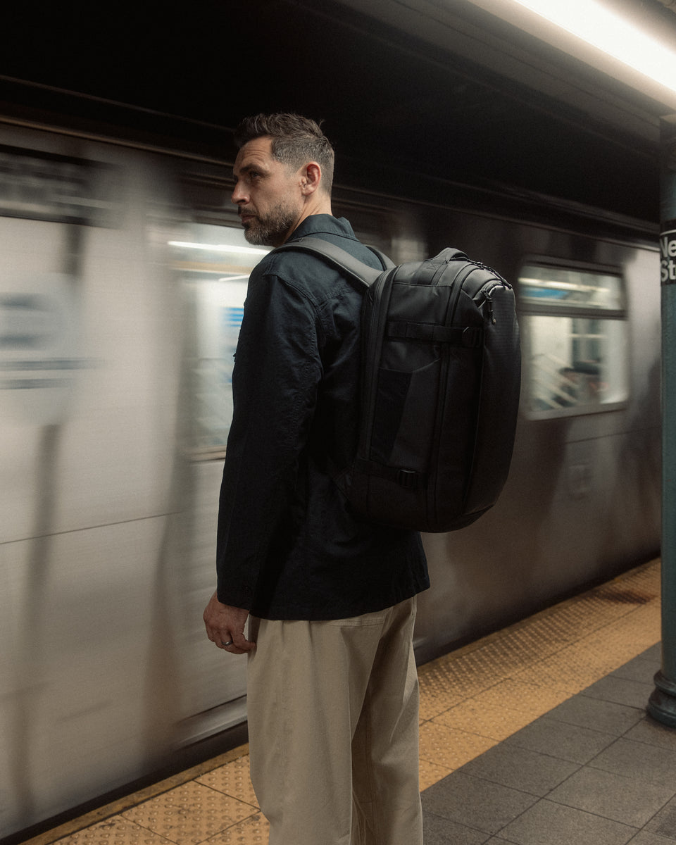 A man standing on a subway platform in New York, standing at an angle wearing a Hybrid Backpack 30L on his back