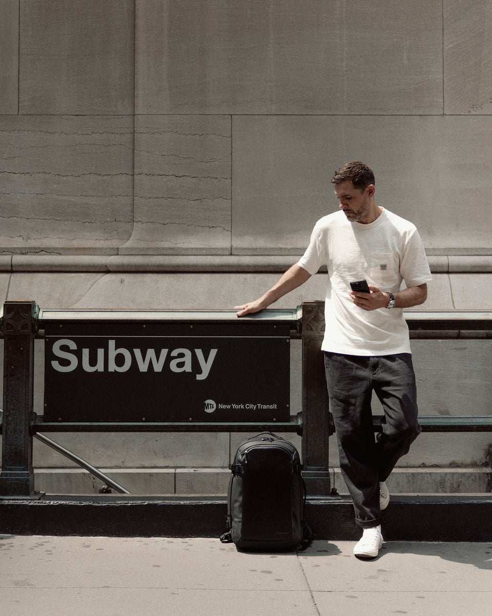 A man standing on his phone next to the Subway entrance in New York with a Hybrid Backpack 30L on the floor next to him