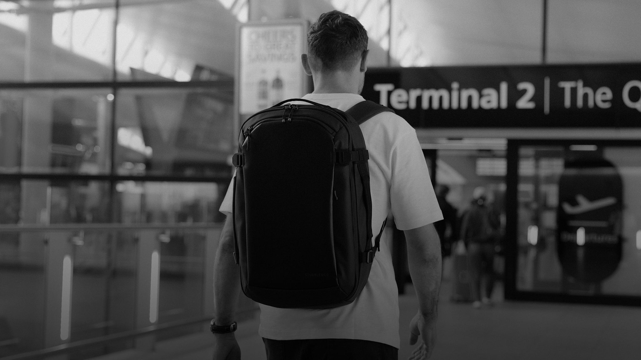 A black and white shot of a man walking into a terminal of an airport with a Hybrid backpack 30l on his back