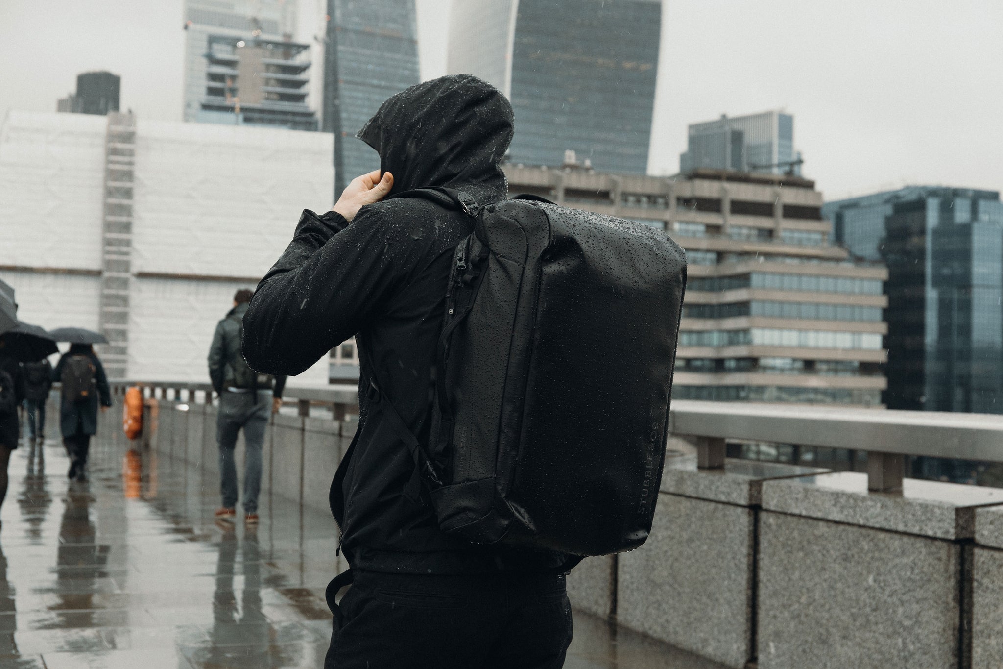 Person in rain coat with hood up wearing a black Kit Bag in the rain