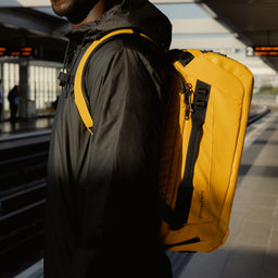 A close up shot of a man wearing a Kit Bag 30L in Yellow on his back whilst standing on a train platform