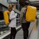 A man and woman standing on an escalator wearing Kit Bags in Yellow on their backs