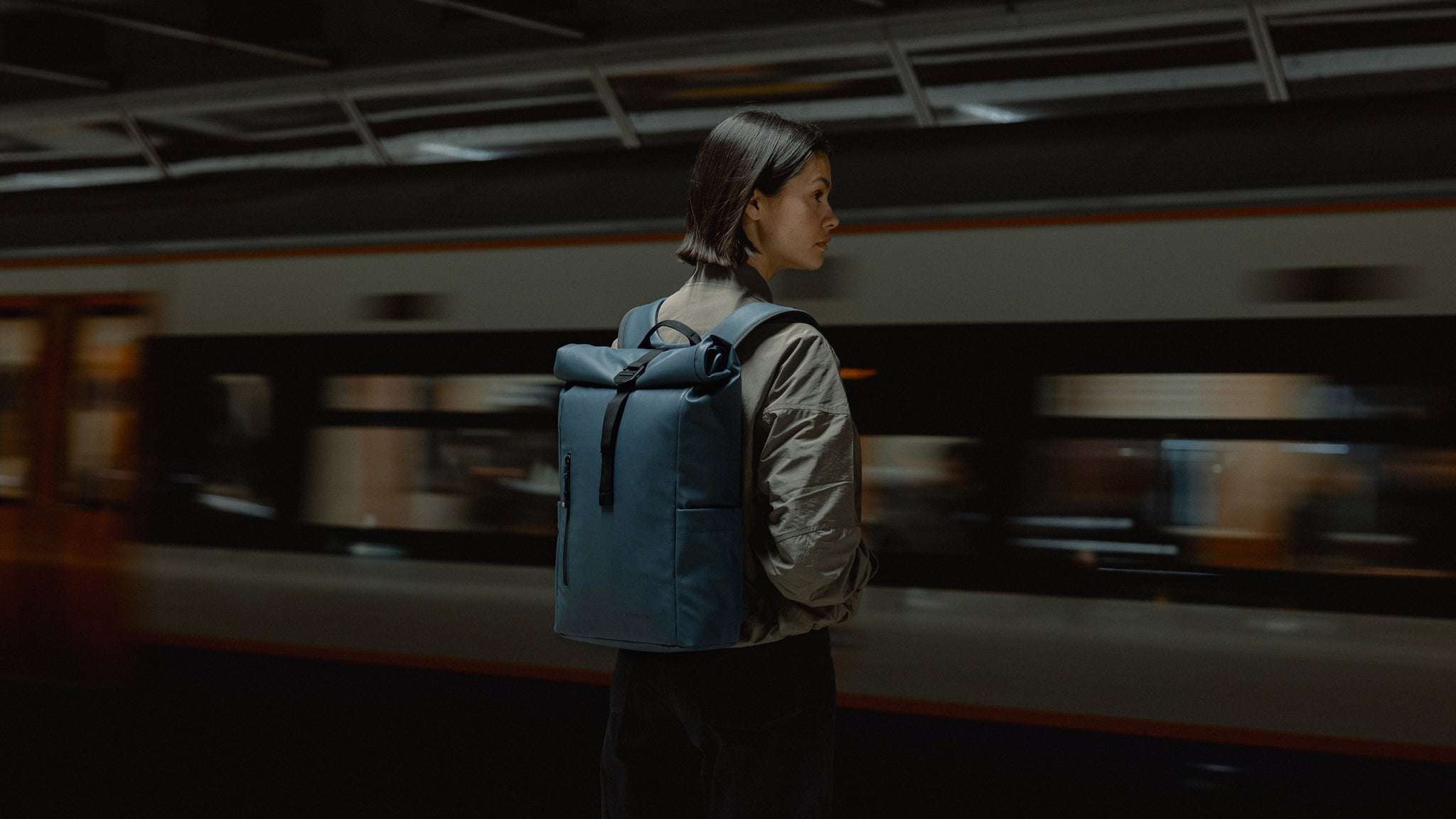 An image of a woman standing on a tube platform as a tube is passing and she is wearing a roll top 15l tamin blue on her back