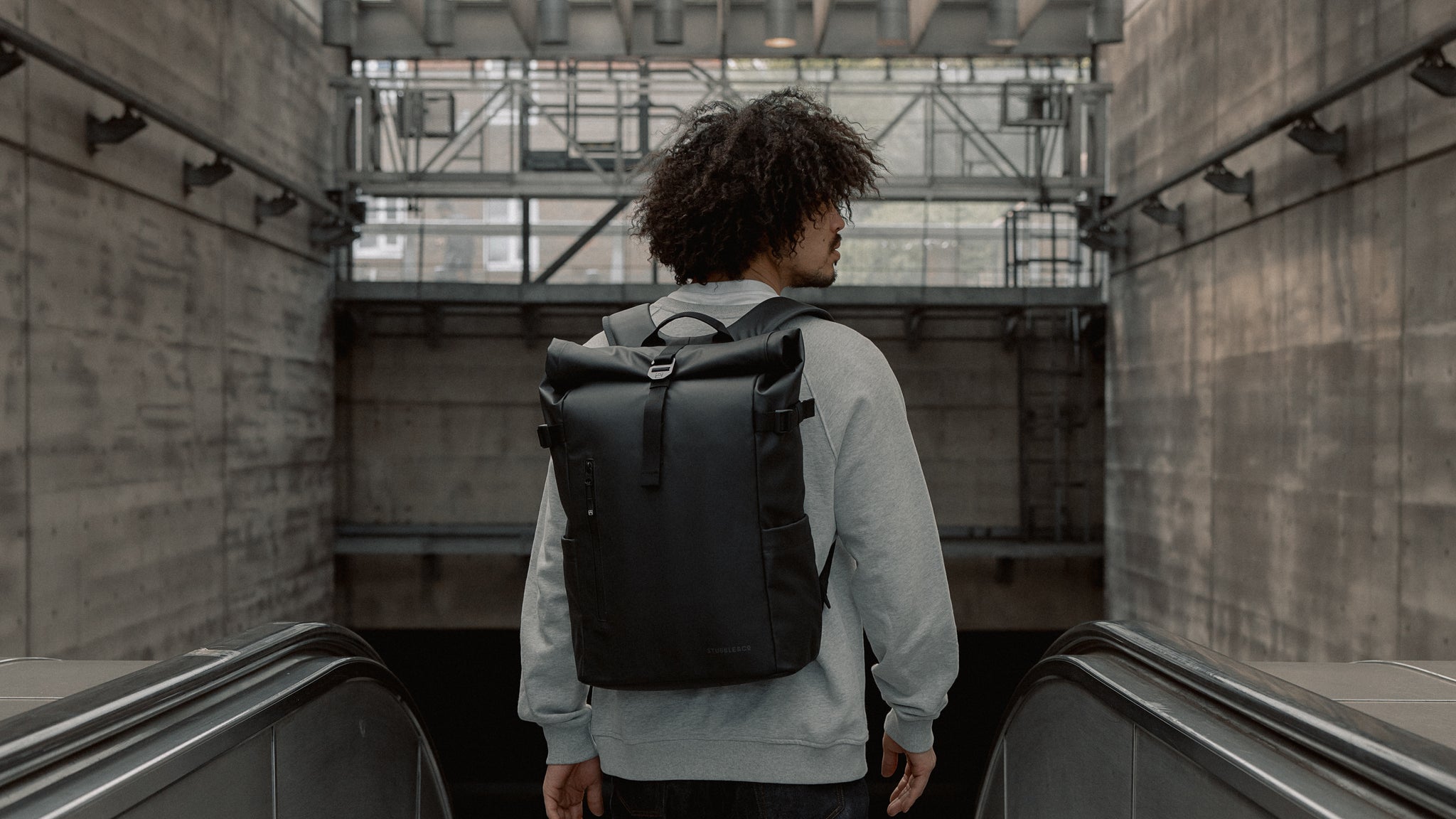 A man standing at the top of the esculators at a tube station with a black roll top 20l on his back