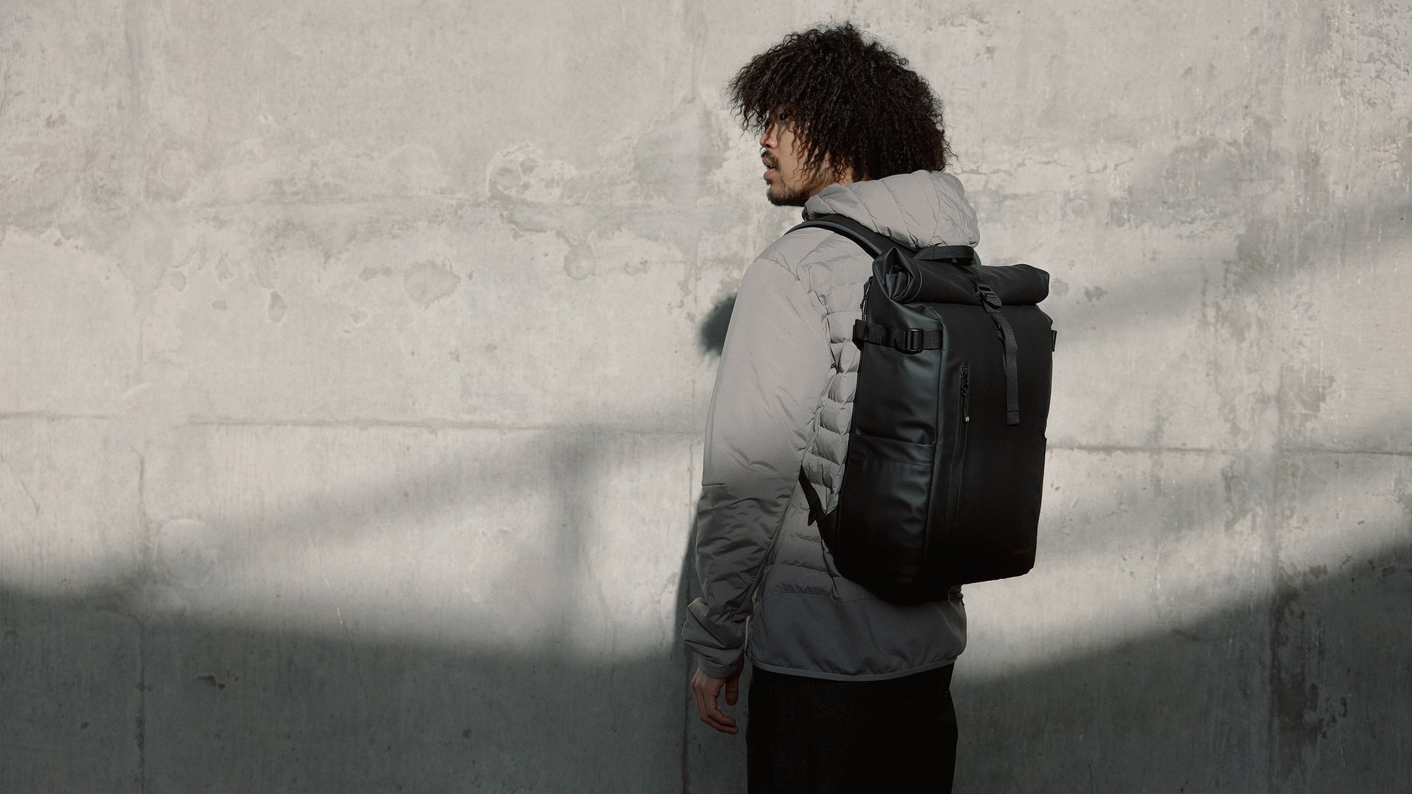 A lifestyle shot of a man wearing a black roll top 20l in front of a concrete wall