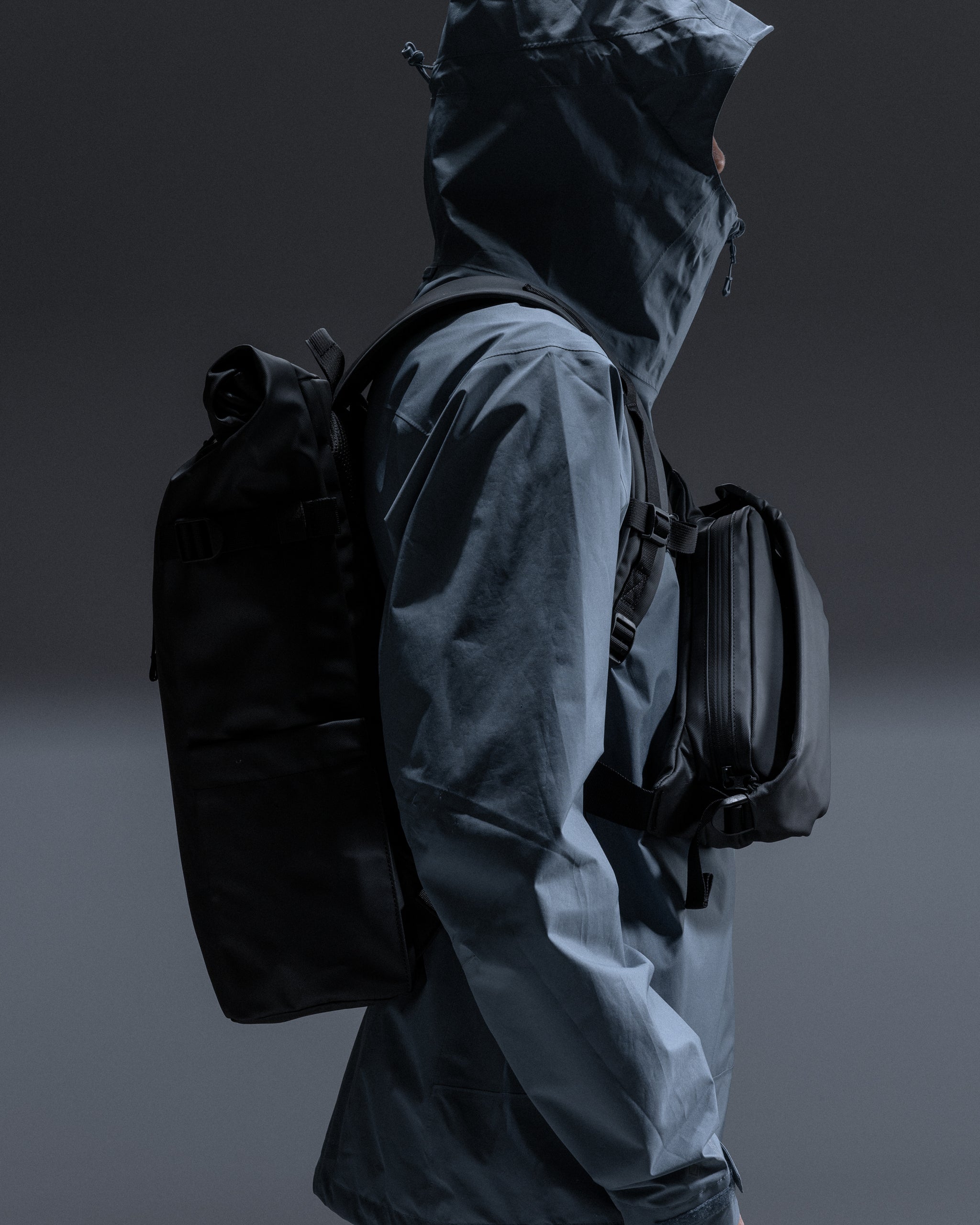 A man wearing a roll top 20l on his back and black crossbody on his front in a studio setting