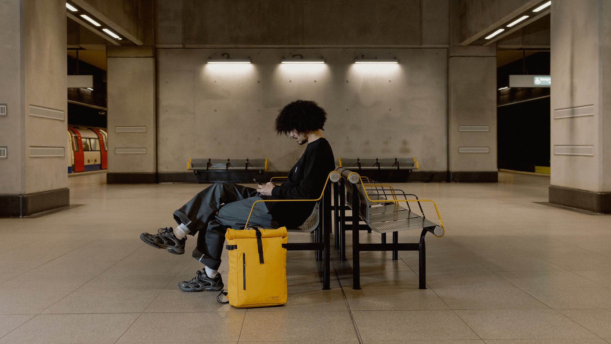 A  man sitting on a bench in a tube station on his phone with a yellow roll top next to him