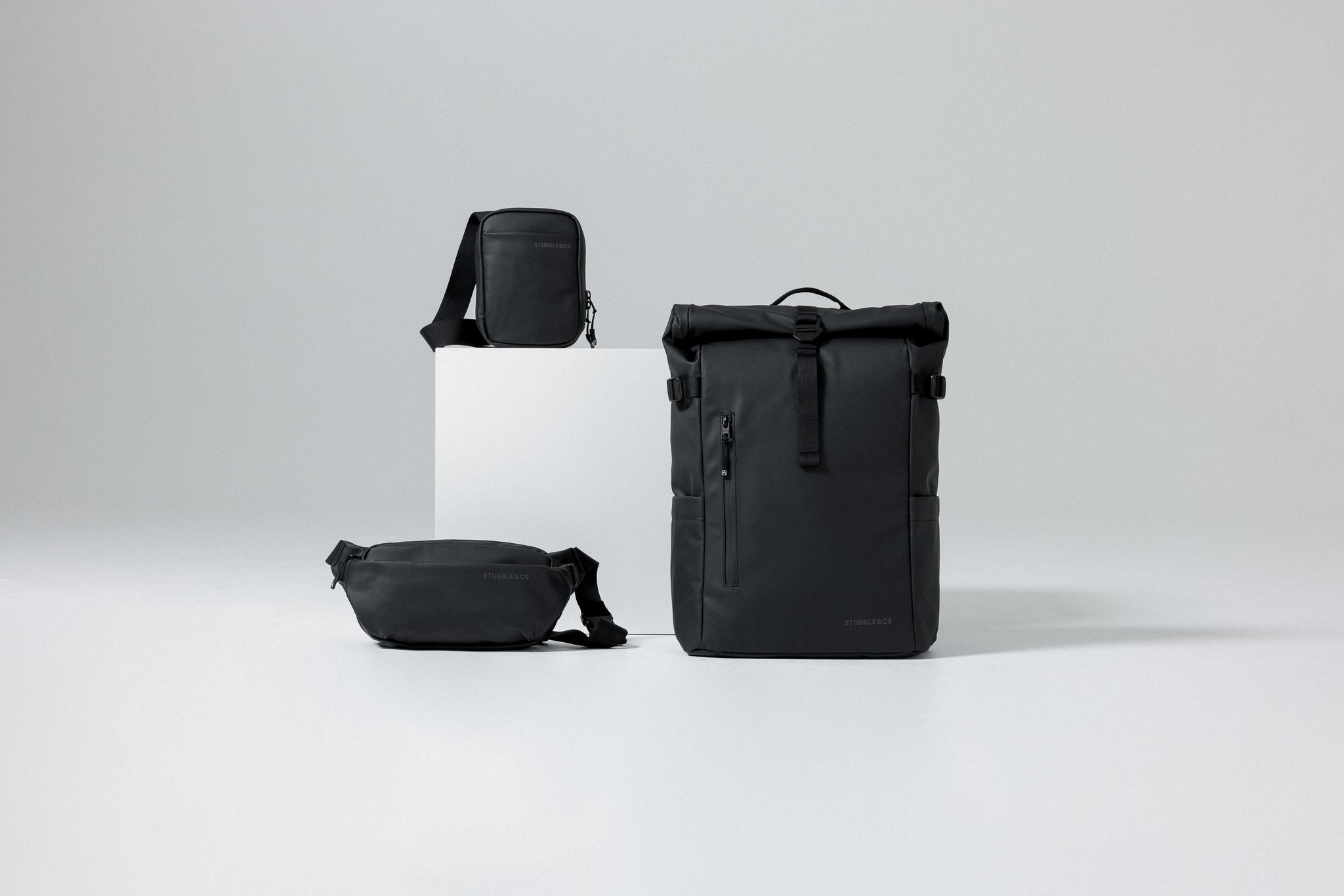 A studio shot of the roll top, shoulder bag and crossbody all in black sitting on plinths.