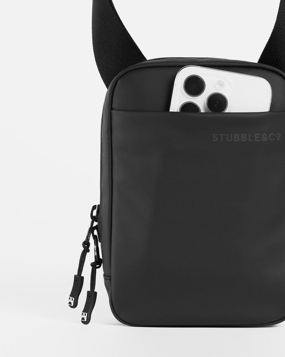 A studio shot of an all black shoulder bag showing the front slip pocket with a phone in it