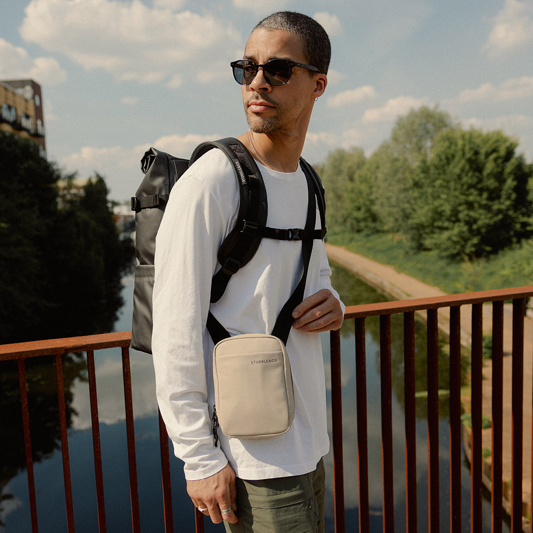 a man standing on a bridge wearing a roll top and Sand shoulder bag