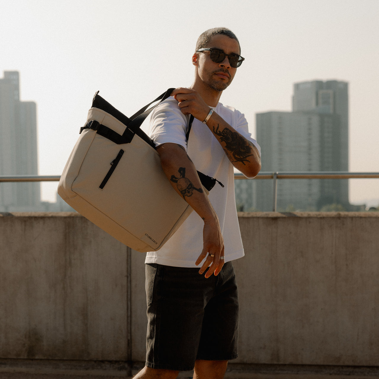 a man putting a Sand Tote Bag onto his shoulder with a city in the background